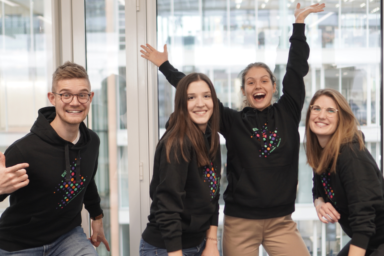 Four Elli designers and engineers excitedly and encouragingly throwing their arms in the air while wearing Elli hoodies at one of Elli's offices.